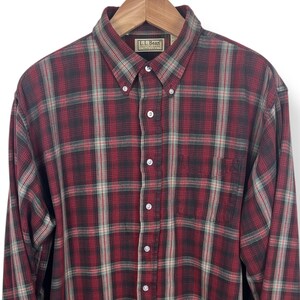 May include: A long-sleeved, button-down plaid shirt in shades of red, black, and green. The shirt has a button-down collar and a chest pocket. The brand label "L.L. Bean" is visible at the top. The shirt is hanging on a wooden hanger.
