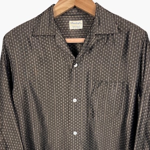 May include: A brown button-down shirt with a small square pattern and vertical stripes. The shirt has a collar, a chest pocket, and long sleeves with buttoned cuffs. The shirt is hanging on a wooden hanger.