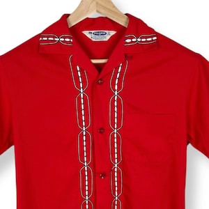 May include: A bright red short-sleeved shirt with a camp collar. The shirt features white and black chain-like detailing down the front and around the collar. A small pocket is visible on the left chest.