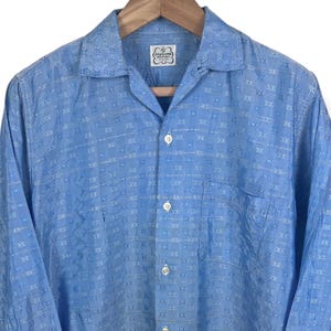 May include: A light blue long-sleeve shirt with a geometric pattern. The shirt has a camp collar, a chest pocket, and buttoned cuffs. The shirt is hanging on a wooden hanger.
