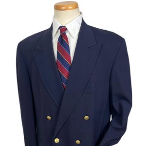 Könnte beinhalten: Ein marineblauer, zweireihiger Blazer mit goldenen Knöpfen, einem weißen Hemd und einer gestreiften Krawatte. Der Blazer hat ein klassisches Design mit fallendem Revers und zwei Vordertaschen. Die Krawatte hat rote, blaue und gelbe Streifen.