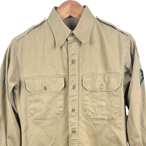 Może przedstawiać: Beżowa koszula z długim rękawem z dwiema kieszeniami na piersi i zapięciem na guziki. Koszula ma kołnierzyk i pagony na ramionach. Mała naszywka jest widoczna na lewym rękawie. Wisi na drewnianym wieszaku.