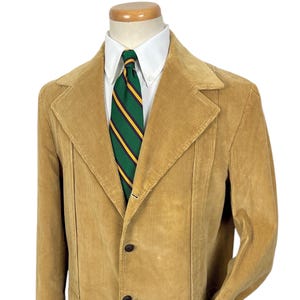 Può includere: Blazer di velluto a coste color cammello con revers a lancia, due bottoni e due tasche anteriori. Il blazer è indossato sopra una camicia bianca con colletto e una cravatta a righe verdi e oro. La giacca è esposta su un manichino.