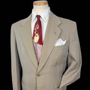 Könnte beinhalten: Beige Anzugjacke mit weißem Hemd und gemusterter Krawatte. Die Jacke hat ein Einstecktuch und zwei Vordertaschen. Der Anzug wird auf einer Schaufensterpuppe präsentiert.