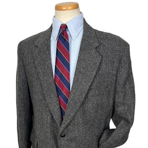 Puede incluir: Una blazer gris con diseño de espiga, camisa azul claro y corbata a rayas rojas, azul marino y doradas. La blazer tiene cierre de un botón y solapa clásica con muesca. El conjunto se exhibe en un maniquí.