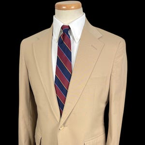 May include: A tan blazer with a two-button closure, a notched lapel, and two front pockets. The blazer is paired with a white dress shirt and a striped tie in navy, burgundy, and gold. The blazer is displayed on a mannequin.