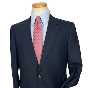 Puede incluir: Un blazer azul marino con una camisa azul claro y una corbata rosa con lunares azul oscuro. El blazer tiene un solo botón dorado y bolsillos. El conjunto se muestra en un maniquí.