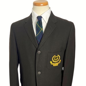Puede incluir: Una blazer marrón oscuro con una camisa de cuello blanco y una corbata a cuadros. La blazer tiene un parche con letras doradas que dice "NEW JERSEY STATE COLLEGE". La blazer tiene dos bolsillos delanteros y tres botones.