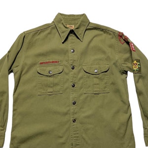 May include: A green long-sleeved button-down shirt with two chest pockets and embroidered patches on the sleeves. The patches feature the words "Boy Scouts of America" and a scout emblem.