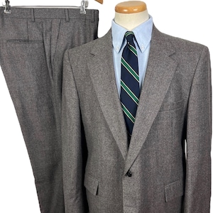 May include: A gray suit with a matching jacket and pants. The jacket has two front pockets and a single-button closure. A light blue collared shirt and a striped tie are visible. The pants are hanging on a wooden hanger.