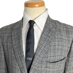 Op de afbeelding: Een grijs en zwart geruit blazer met een zwarte stropdas. De blazer heeft een enkele knoopsluiting en een zak aan de linkerkant.