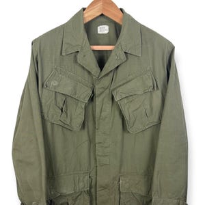 Może przedstawiać: Zielona kurtka w stylu wojskowym z czterema kieszeniami z przodu i kołnierzem. Kurtka wisi na drewnianym wieszaku na białym tle. Ma zapięcie na guziki i długie rękawy.