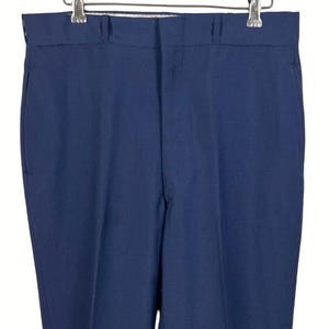 May include: A pair of solid navy blue trousers featuring a structured waistband with belt loops, a front zipper fly closure, and a clean, straight-leg cut. The pants are displayed hanging from a wooden hanger with metal clips.