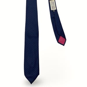May include: A dark blue necktie featuring a pattern of tiny red dots. The tie has a pointed end and a label on the back that reads Bonwit Teller Mens Shop. The underside of the tip is lined with a solid pink fabric, providing a contrast to the blue.