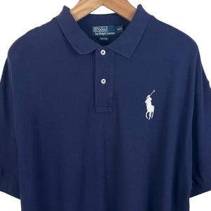 Puede incluir: Un polo azul marino con mangas cortas, cuello y tapeta de tres botones. La camisa presenta un logotipo blanco bordado de un jugador de polo en el pecho izquierdo. La camisa está colgada de una percha de madera.