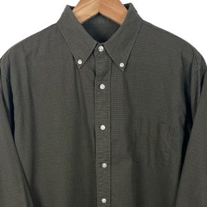 May include: A dark olive green button-down shirt, displayed on a wooden hanger. The shirt has a small check pattern and a chest pocket. It features a button-down collar and white buttons.
