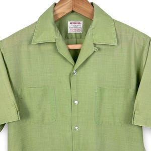 May include: A light green short-sleeve button-down shirt with two chest pockets. The shirt has a camp collar and is hanging on a wooden hanger. The brand tag reads "McGregor SCOTSER".