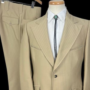 May include: A tan suit with a matching jacket and pants, displayed on a mannequin. The jacket features a two-button closure, notched lapels, and flap pockets. A white dress shirt and bolo tie with a turquoise stone complete the ensemble. The pants are hanging on a hanger.