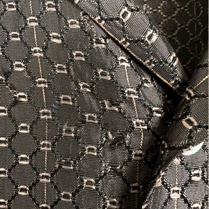 May include: Close-up of a black and silver patterned fabric, possibly from a blazer or jacket. The fabric has a repeating geometric design with small, rectangular silver accents. The texture appears to be woven, with a subtle sheen.