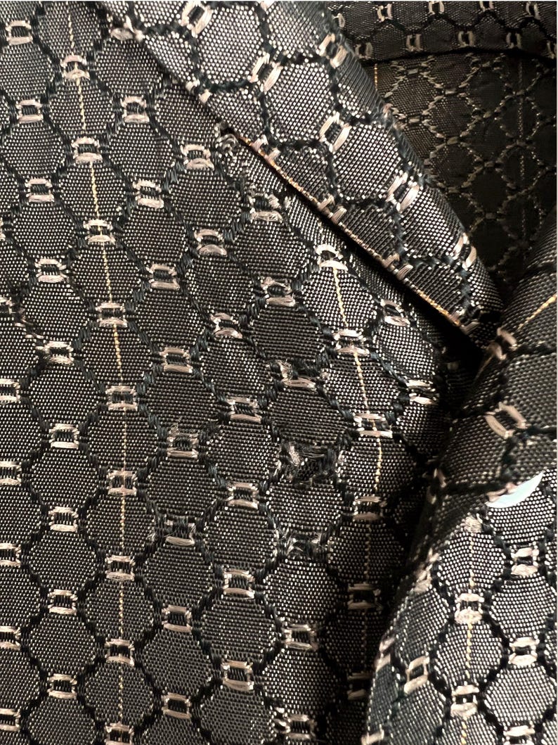 May include: Close-up of a black and silver patterned fabric, possibly from a blazer or jacket. The fabric has a repeating geometric design with small, rectangular silver accents. The texture appears to be woven, with a subtle sheen.