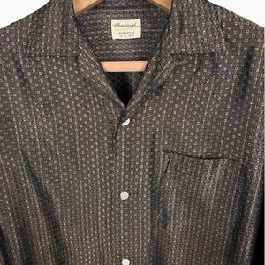 May include: A brown button-down shirt with a geometric pattern, hanging on a wooden hanger. The shirt has a camp collar, a chest pocket, and white buttons. The tag reads "Pennleigh Washable".