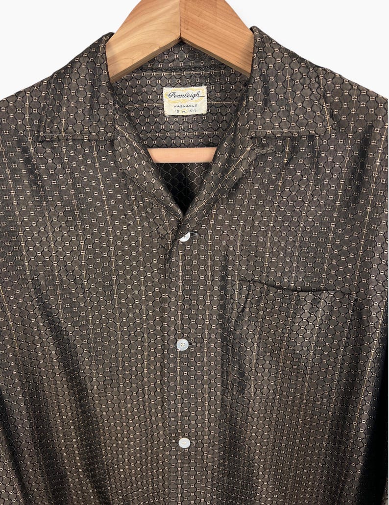 May include: A brown button-down shirt with a geometric pattern, hanging on a wooden hanger. The shirt has a camp collar, a chest pocket, and white buttons. The tag reads "Pennleigh Washable".