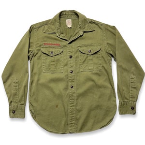 May include: A green long-sleeved button-up shirt with two large pockets and a red embroidered patch that says "Boy Scouts of America".