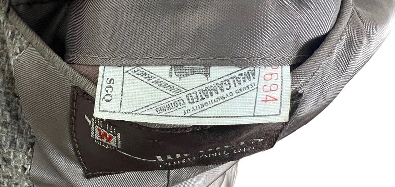 May include: A white tag with black text that reads "Issued by authority of " followed by a logo and the text "Mallalieu Limited" and "John Ford & Son" with a red number "2694" on the right side. The tag is attached to a brown fabric with a white tag that reads "SCQ" in black text.