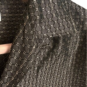 May include: Close-up of a dark gray blazer with a repeating geometric pattern. The fabric has a textured appearance with a grid-like design, featuring small, light-colored accents. The blazer is hanging on a wooden hanger.