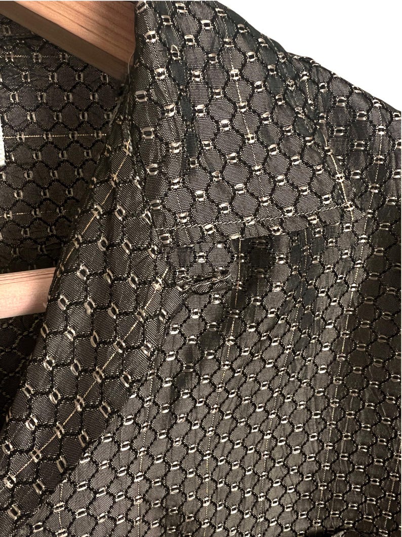 May include: Close-up of a dark gray blazer with a repeating geometric pattern. The fabric has a textured appearance with a grid-like design, featuring small, light-colored accents. The blazer is hanging on a wooden hanger.