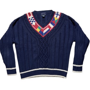 May include: A navy blue cable knit sweater with a colorful nautical flag pattern on the v-neck. The sweater has white trim on the cuffs and hem.