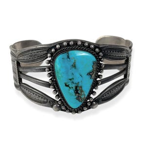 May include: A silver cuff bracelet featuring a large, turquoise stone. The stone is a bright blue-green with dark brown markings. The bracelet has an engraved design and is a striking piece of jewellery.
