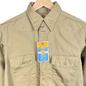 May include: A tan long-sleeve shirt with a button-down collar and two chest pockets. The shirt has a label that reads "Biltowell Shirt Raw Hide Sanforized".