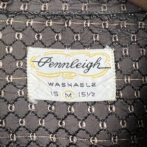 May include: Close-up of a vintage shirt label with the brand name "Pennleigh" in cursive script. The label is white with a gold border and the word "Washable" is printed below. The shirt fabric has a geometric pattern in black and tan.