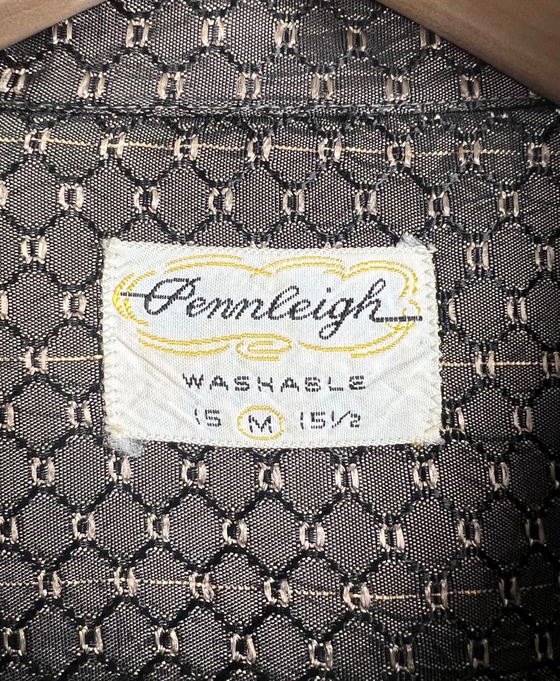 May include: Close-up of a vintage shirt label with the brand name "Pennleigh" in cursive script. The label is white with a gold border and the word "Washable" is printed below. The shirt fabric has a geometric pattern in black and tan.