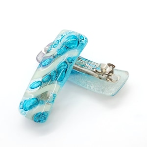 May include: Two rectangular hair clips with a glass-like appearance. The clips feature a design of turquoise and white swirls and shapes, with a silver metal clasp. The clips are set against a white background.