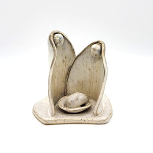 Puede incluir: Un belén de cerámica con acabado color crema. La escultura presenta dos figuras que flanquean un pequeño pesebre. Las figuras tienen cabezas redondeadas y están sobre una base moteada. El diseño es minimalista y artístico.