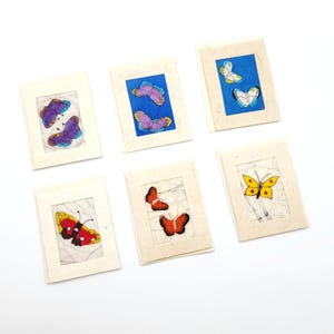 May include: Six rectangular greeting cards with off-white textured borders. Each card features a central illustration of butterflies in various colors including purple, blue, white, yellow, red, and orange, set against different background patterns.
