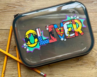 May include: A clear pencil case with the name "OLIVER" in colorful superhero-themed letters. The case is rectangular with a black zipper and is surrounded by yellow pencils.