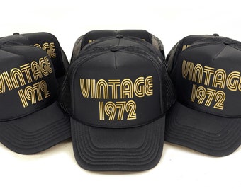 Könnte beinhalten: Drei schwarze Trucker-Caps mit goldenen Netzrücken und goldener Aufschrift "Vintage 1972".
