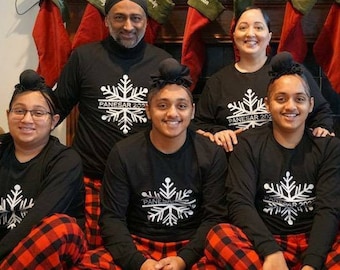 May include: A family of six wearing matching black pajamas with a white snowflake design and the text "PANESAR 2022". They are posing in front of a fireplace decorated with Christmas stockings.