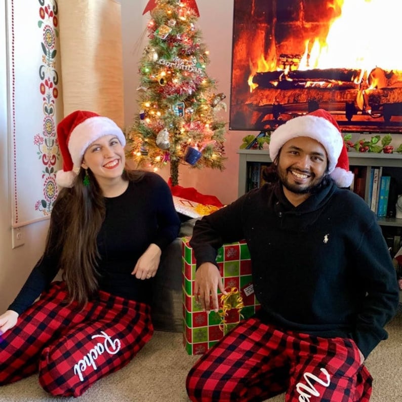 K&ouml;nnte beinhalten: Ein Paar tr&auml;gt passende rote und schwarze karierte Schlafanz&uuml;ge mit den Stickereien "Papa" und "Mama". Sie tragen Weihnachtsm&uuml;tzen und l&auml;cheln in die Kamera. Im Hintergrund steht ein Weihnachtsbaum.