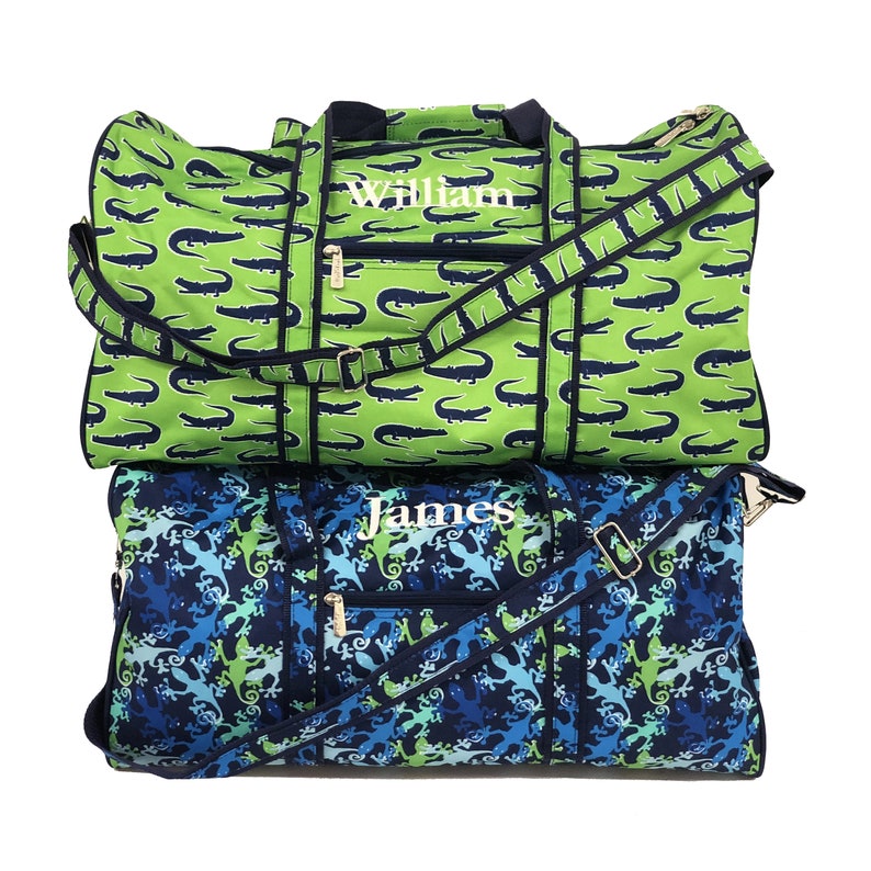 Pu&ograve; includere: Due borse da viaggio personalizzate con stampa di alligatore verde e stampa di lucertola blu e verde. La borsa superiore ha il nome "William" ricamato e la borsa inferiore ha il nome "James" ricamato.