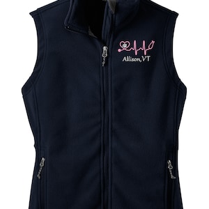 May include: A navy blue fleece vest with a zipper closure and two zippered pockets. The vest has a pink embroidered design of a heart with a stethoscope and paw print, and the text "Allison, VT".