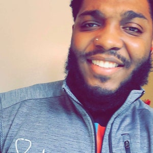 May include: A man with a beard and a nose ring is smiling. He is wearing a gray and white striped jacket with a stethoscope and a heartbeat graphic on the left side. The text "Christopher, RN" is printed below the graphic.