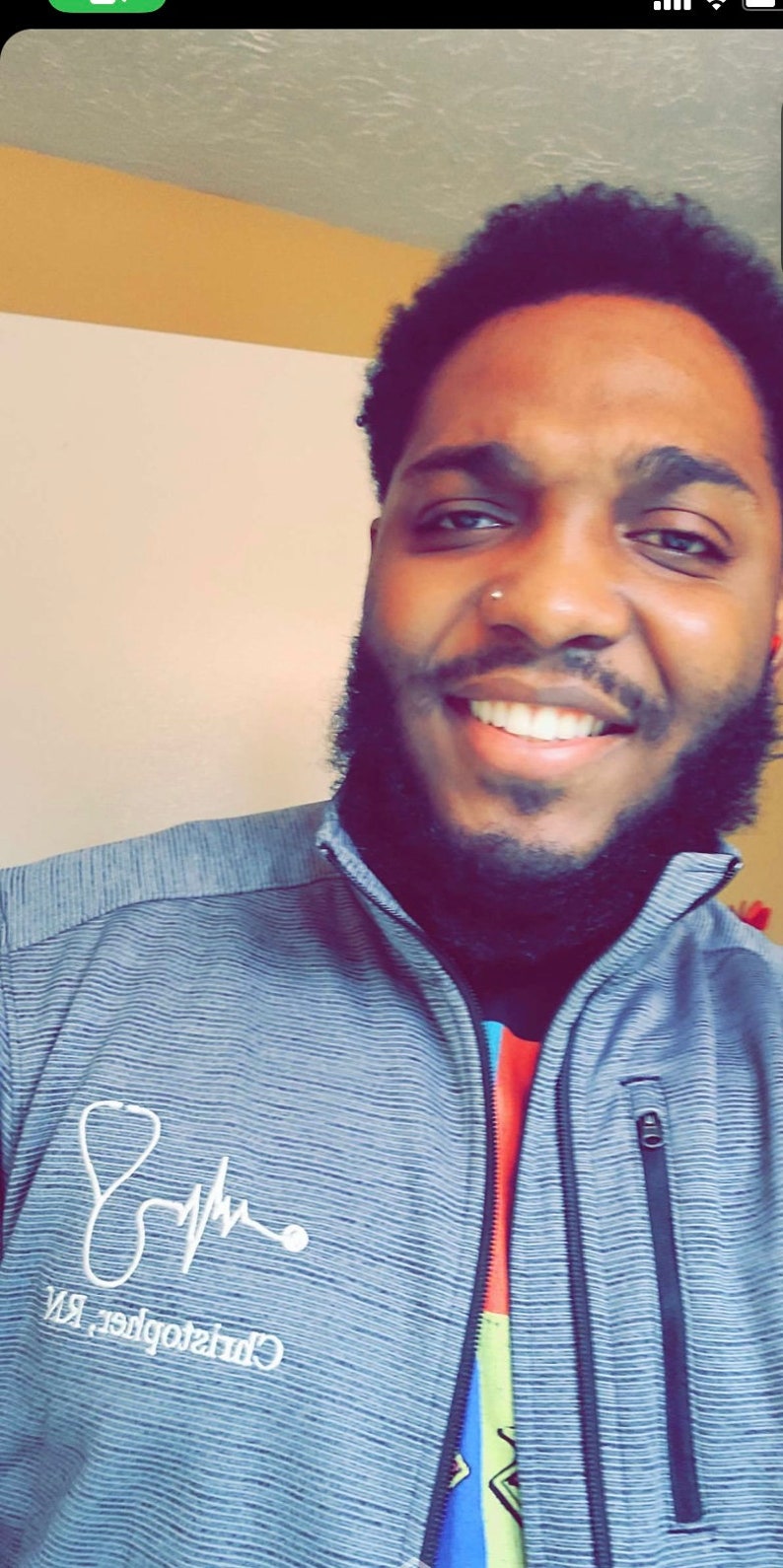 May include: A man with a beard and a nose ring is smiling. He is wearing a gray and white striped jacket with a stethoscope and a heartbeat graphic on the left side. The text "Christopher, RN" is printed below the graphic.