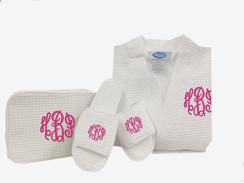 May include: White waffle weave robe, slippers, and a toiletry bag with pink monogram embroidery. The monogram is the letters "HBD".