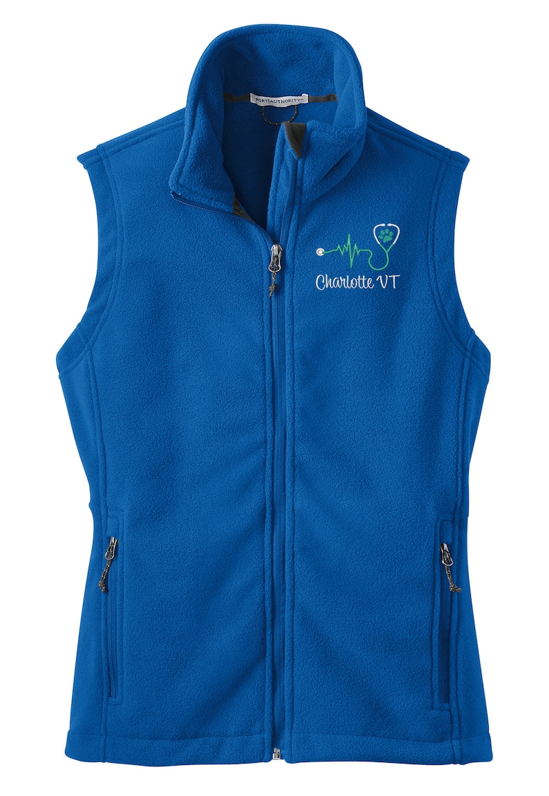 May include: A royal blue fleece vest with a zipper closure. The vest has two zippered pockets on the sides. The vest has an embroidered design on the front that includes a stethoscope, a heartbeat, a paw print, and the text "Charlotte VT".