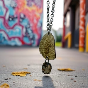 May include: A silver chain necklace with a pendant featuring a large, irregular, amber-colored stone and a smaller, faceted, brown stone. The necklace hangs against a backdrop of colorful graffiti and a paved surface.