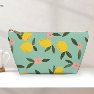 以下が含まれることがあります： レモンと花の模様がついた薄い青色のジッパー付きポーチ。ポーチには金色のジッパーが付いており、ポーチには「vivir furlong」という文字が印刷されています。